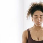 A woman with curly hair meditates peacefully indoors, eyes closed.